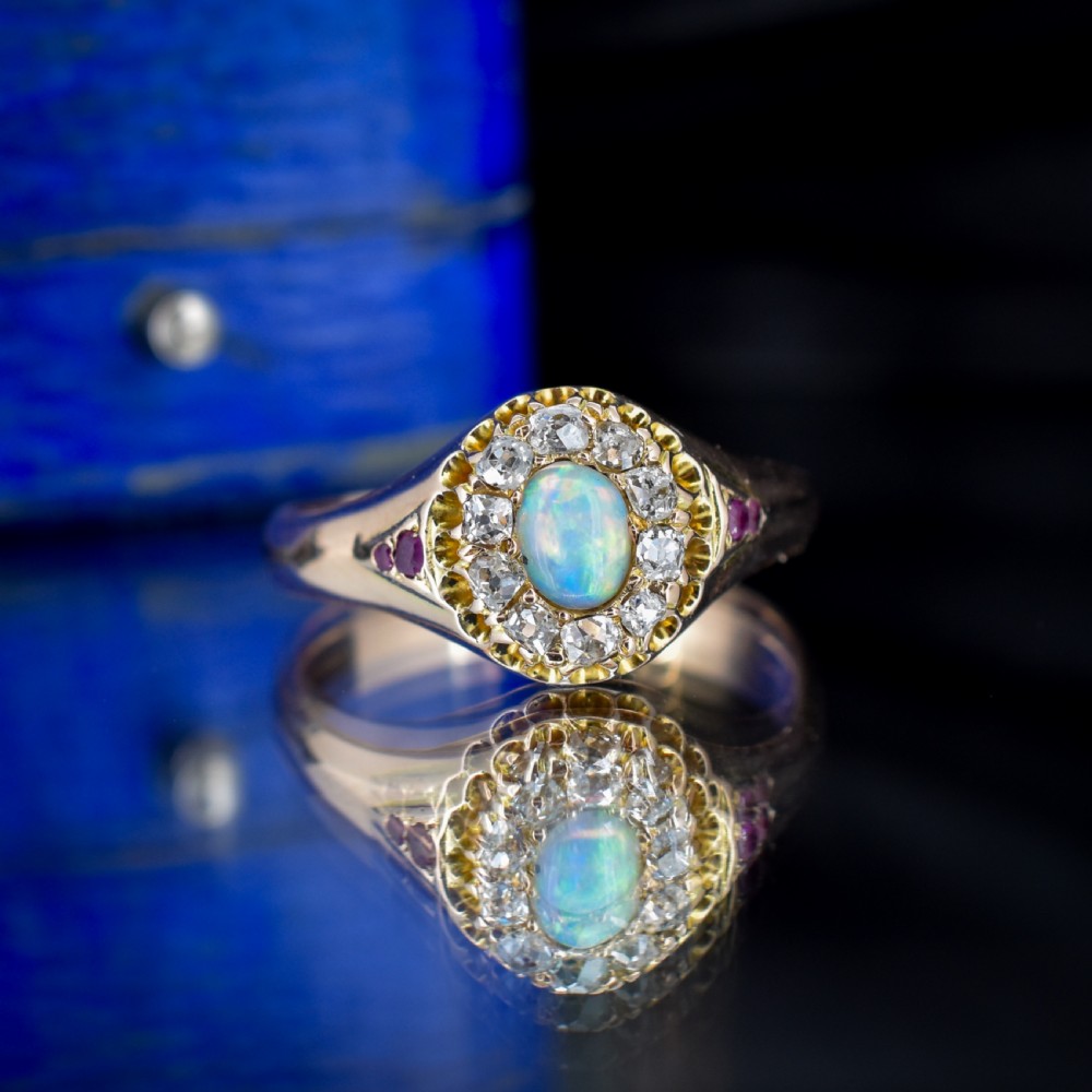 An 18ct yellow gold Victorian ring featuring an oval cabochon opal surrounded by a cluster of diamonds and ruby accents set into the shoulders. View IV