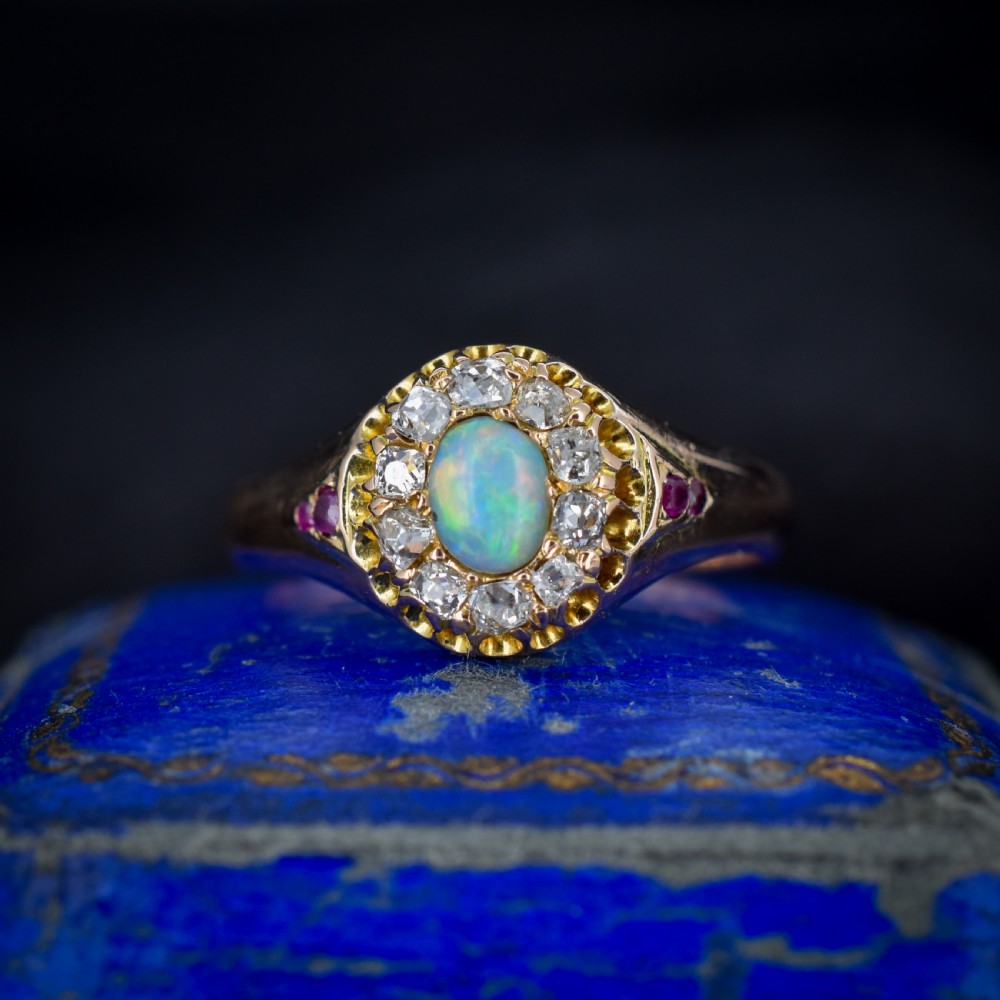 An 18ct yellow gold Victorian ring featuring an oval cabochon opal surrounded by a cluster of diamonds and ruby accents set into the shoulders. View II
