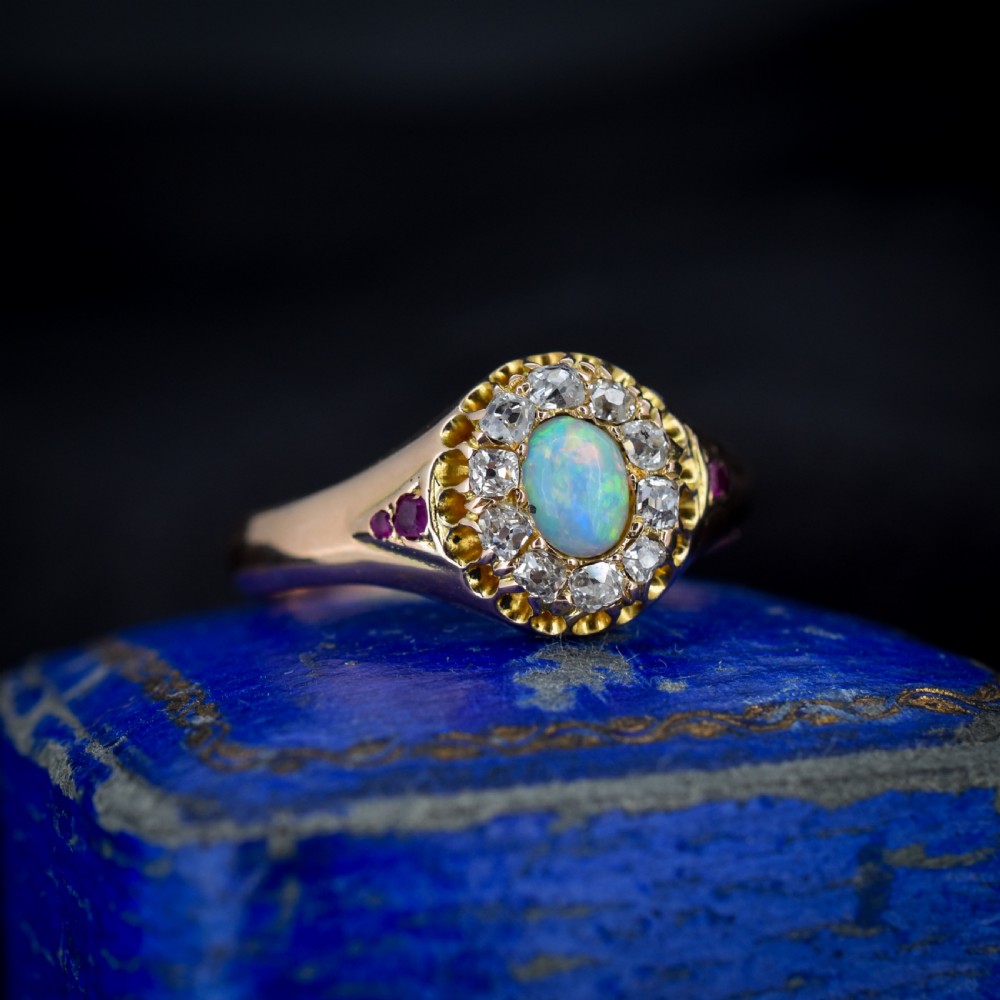An 18ct yellow gold Victorian ring featuring an oval cabochon opal surrounded by a cluster of diamonds and ruby accents set into the shoulders.