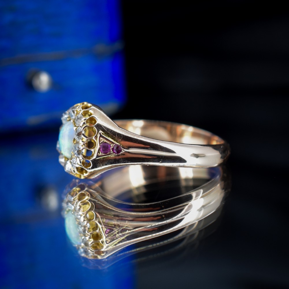 An 18ct yellow gold Victorian ring featuring an oval cabochon opal surrounded by a cluster of diamonds and ruby accents set into the shoulders. View V