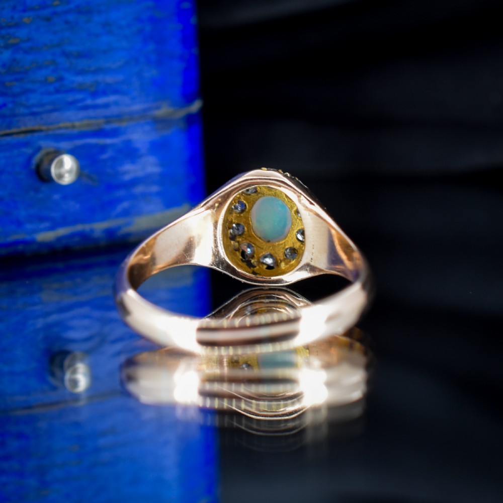 An 18ct yellow gold Victorian ring featuring an oval cabochon opal surrounded by a cluster of diamonds and ruby accents set into the shoulders. View VI