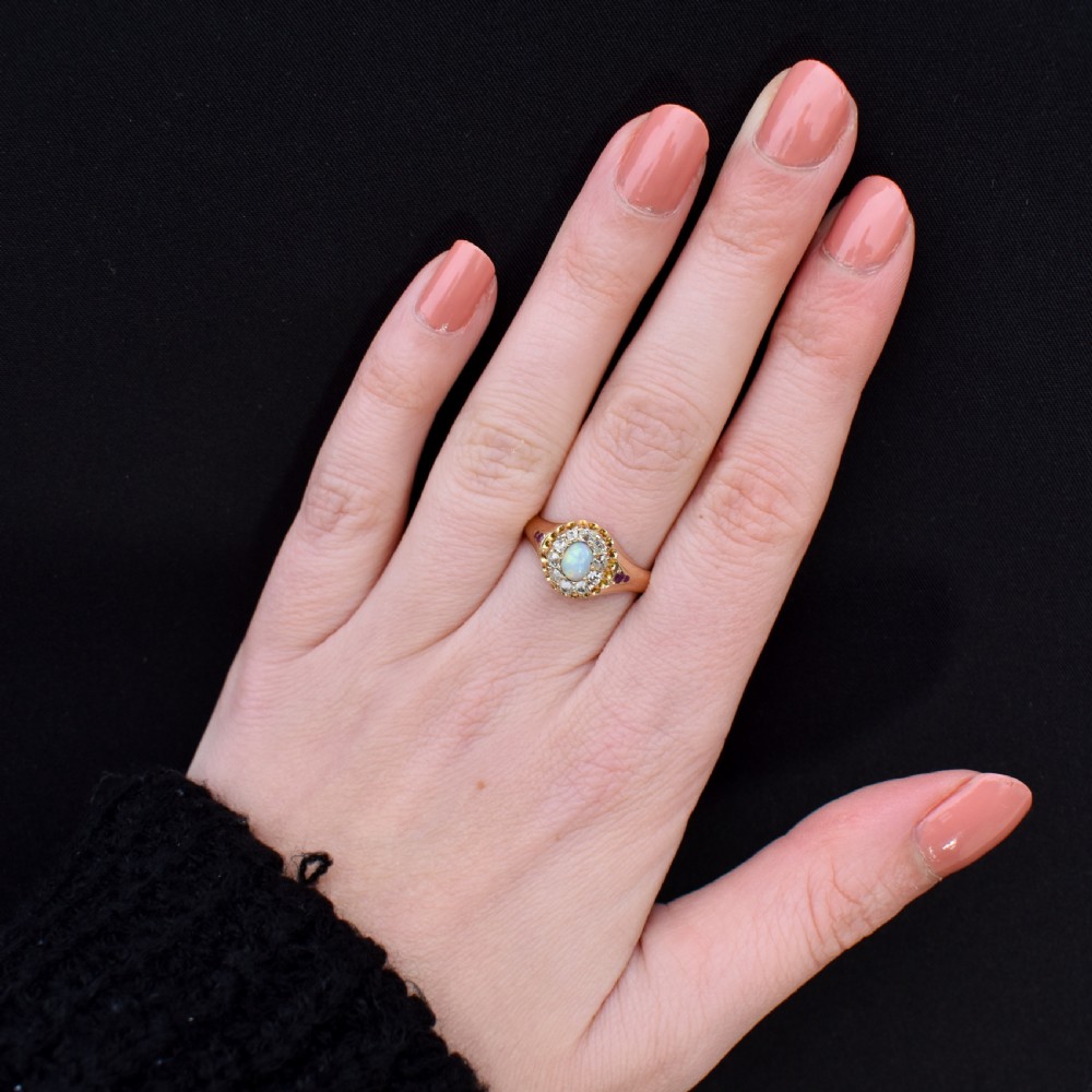 An 18ct yellow gold Victorian ring featuring an oval cabochon opal surrounded by a cluster of diamonds and ruby accents set into the shoulders. View VII