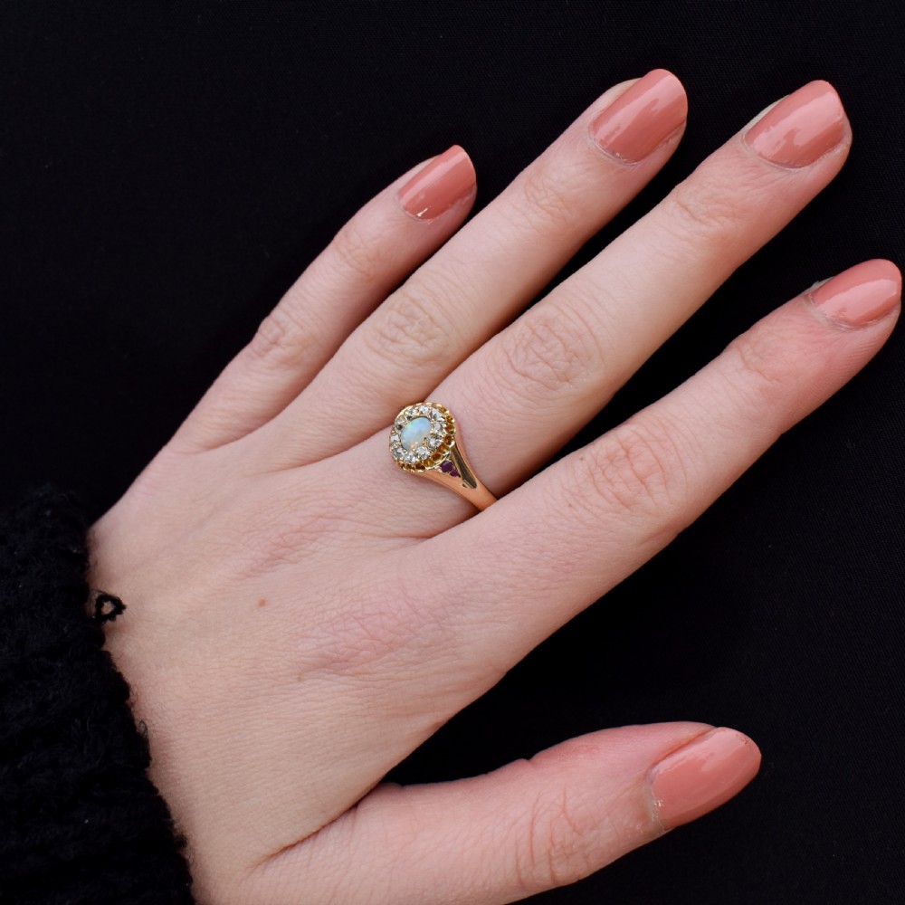 An 18ct yellow gold Victorian ring featuring an oval cabochon opal surrounded by a cluster of diamonds and ruby accents set into the shoulders. View VIII