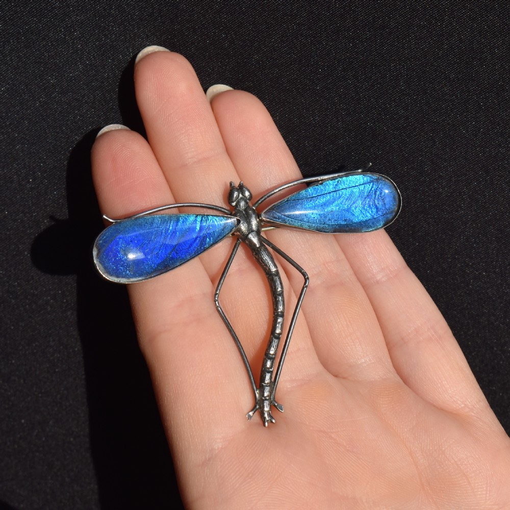 An Art Deco sterling silver brooch designed in the shape of a dragonfly with iridescent blue butterfly wings set beneath glass. View X