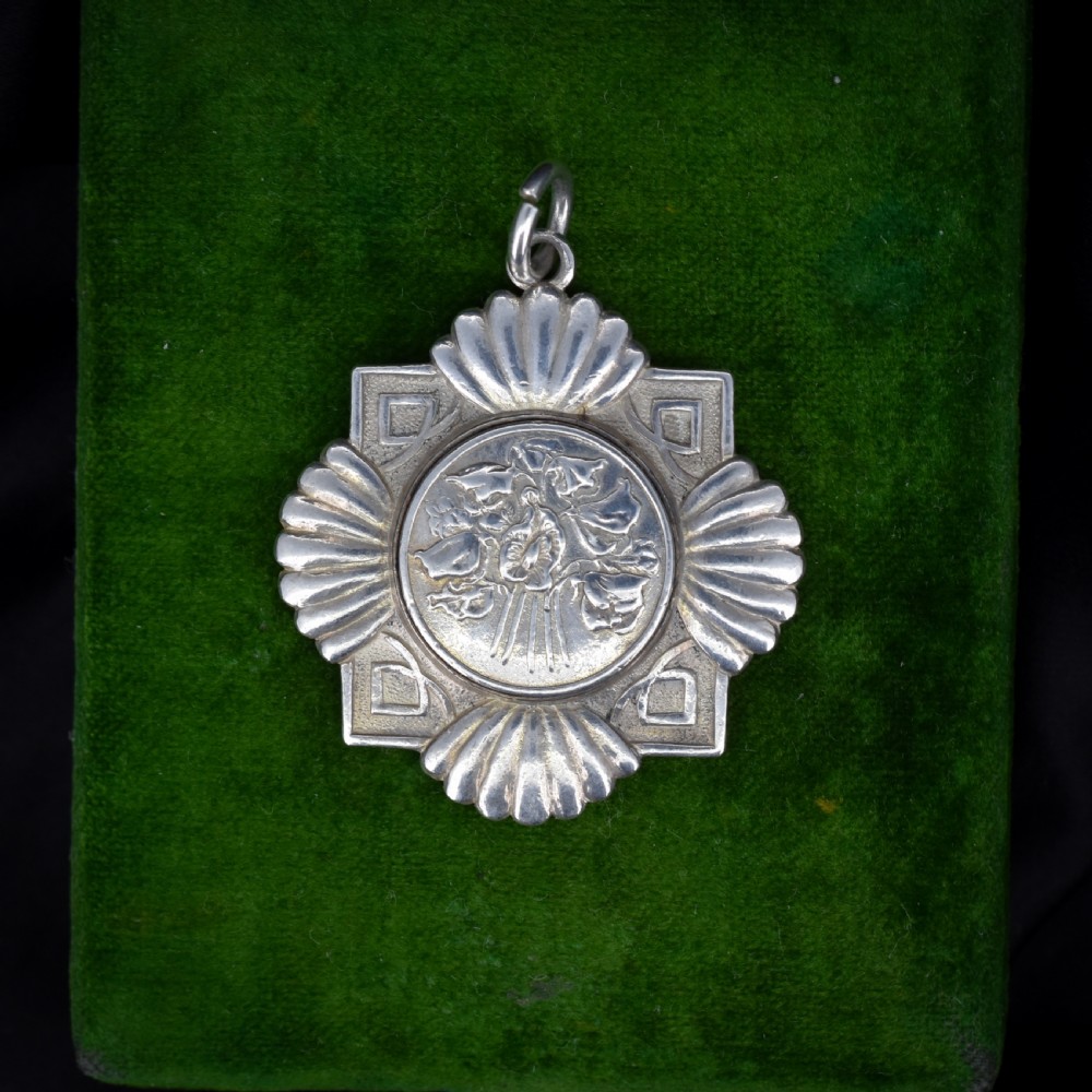 An Art Deco silver medallion featuring a central circular floral relief surrounded by a geometric square and fan-shaped fluted edges with a top suspension loop.