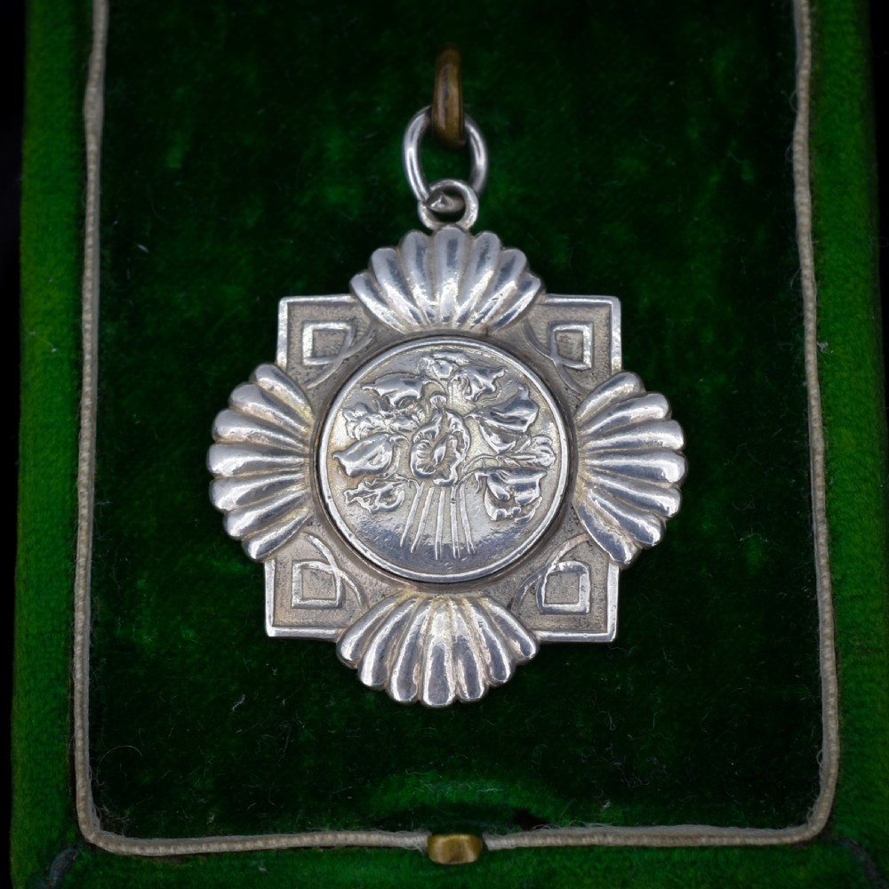 An Art Deco silver medallion featuring a central circular floral relief surrounded by a geometric square and fan-shaped fluted edges with a top suspension loop. View II
