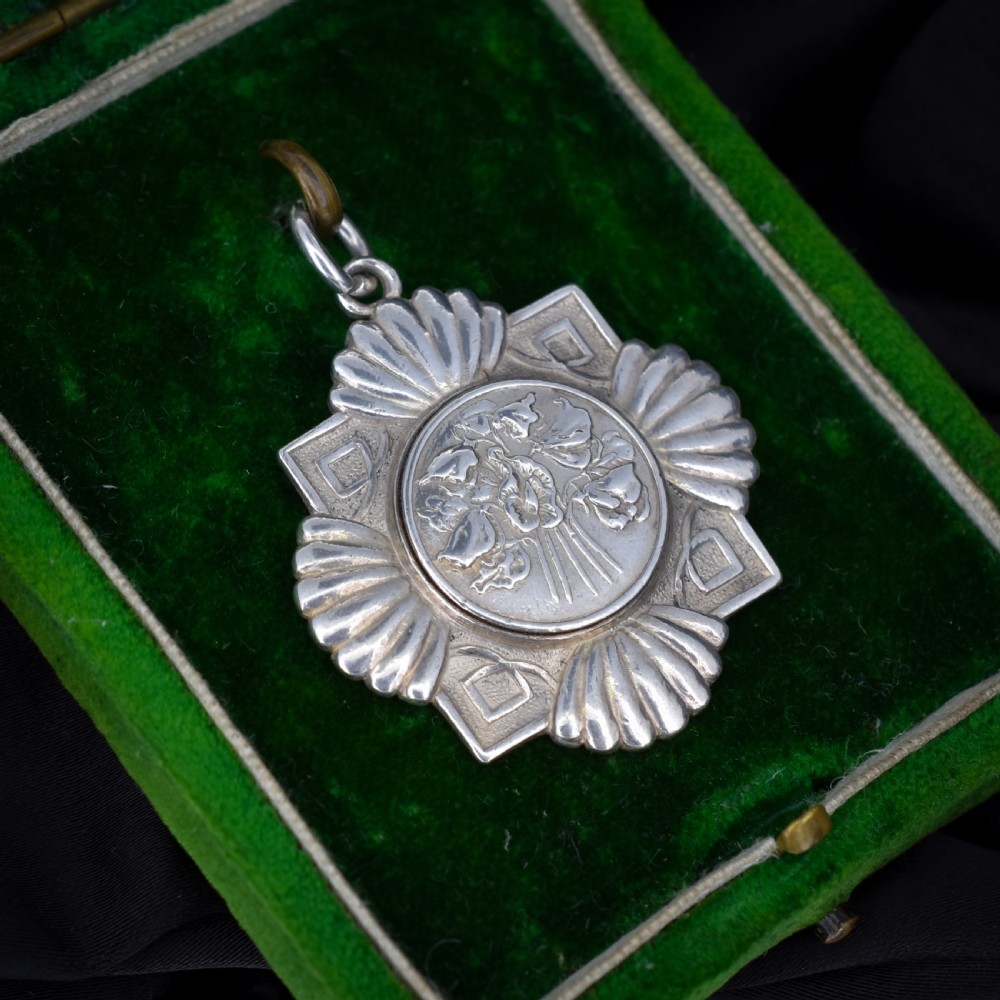 An Art Deco silver medallion featuring a central circular floral relief surrounded by a geometric square and fan-shaped fluted edges with a top suspension loop. View III