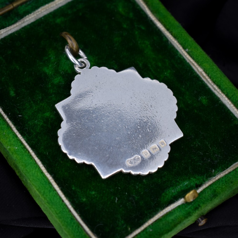 An Art Deco silver medallion featuring a central circular floral relief surrounded by a geometric square and fan-shaped fluted edges with a top suspension loop. View IV