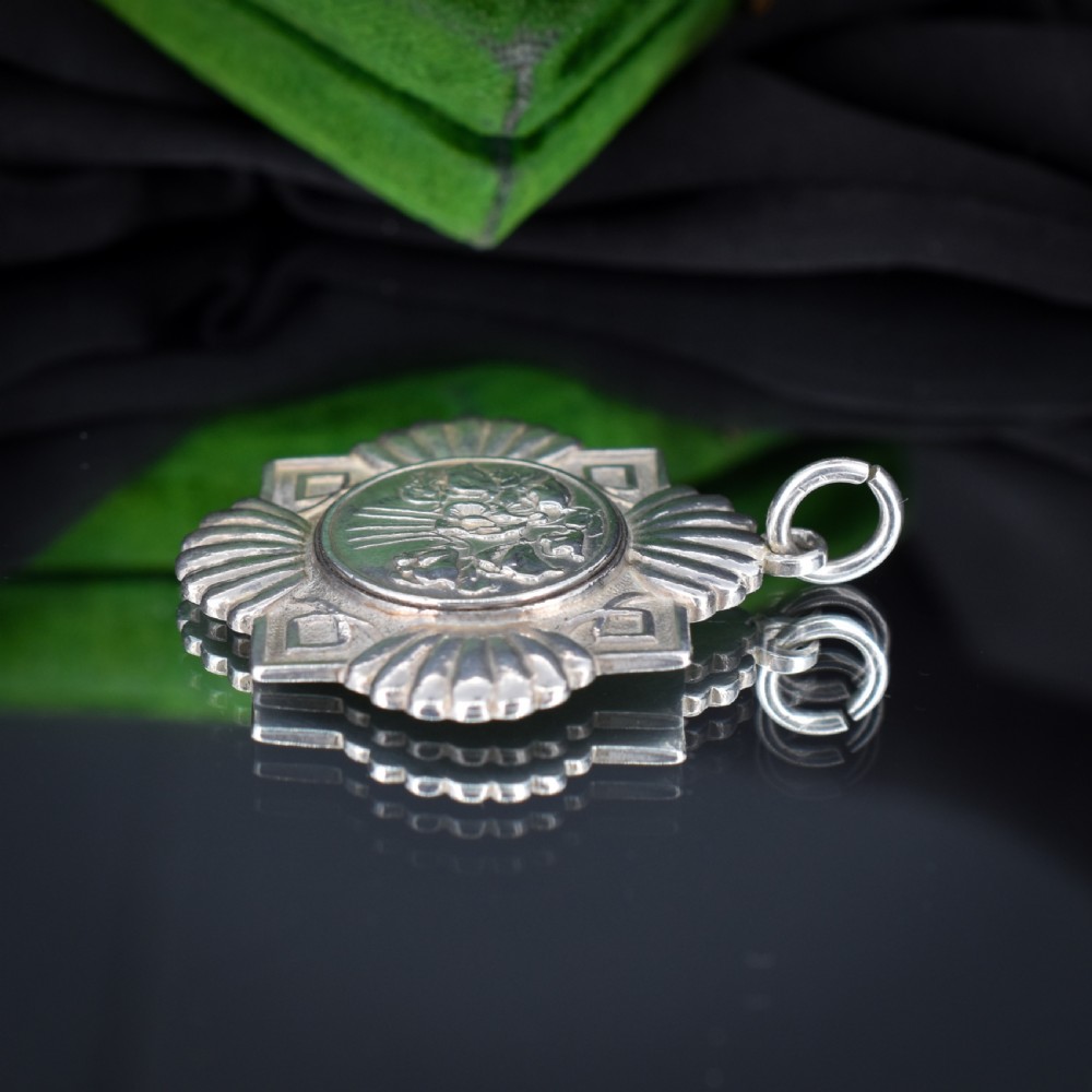 An Art Deco silver medallion featuring a central circular floral relief surrounded by a geometric square and fan-shaped fluted edges with a top suspension loop. View V