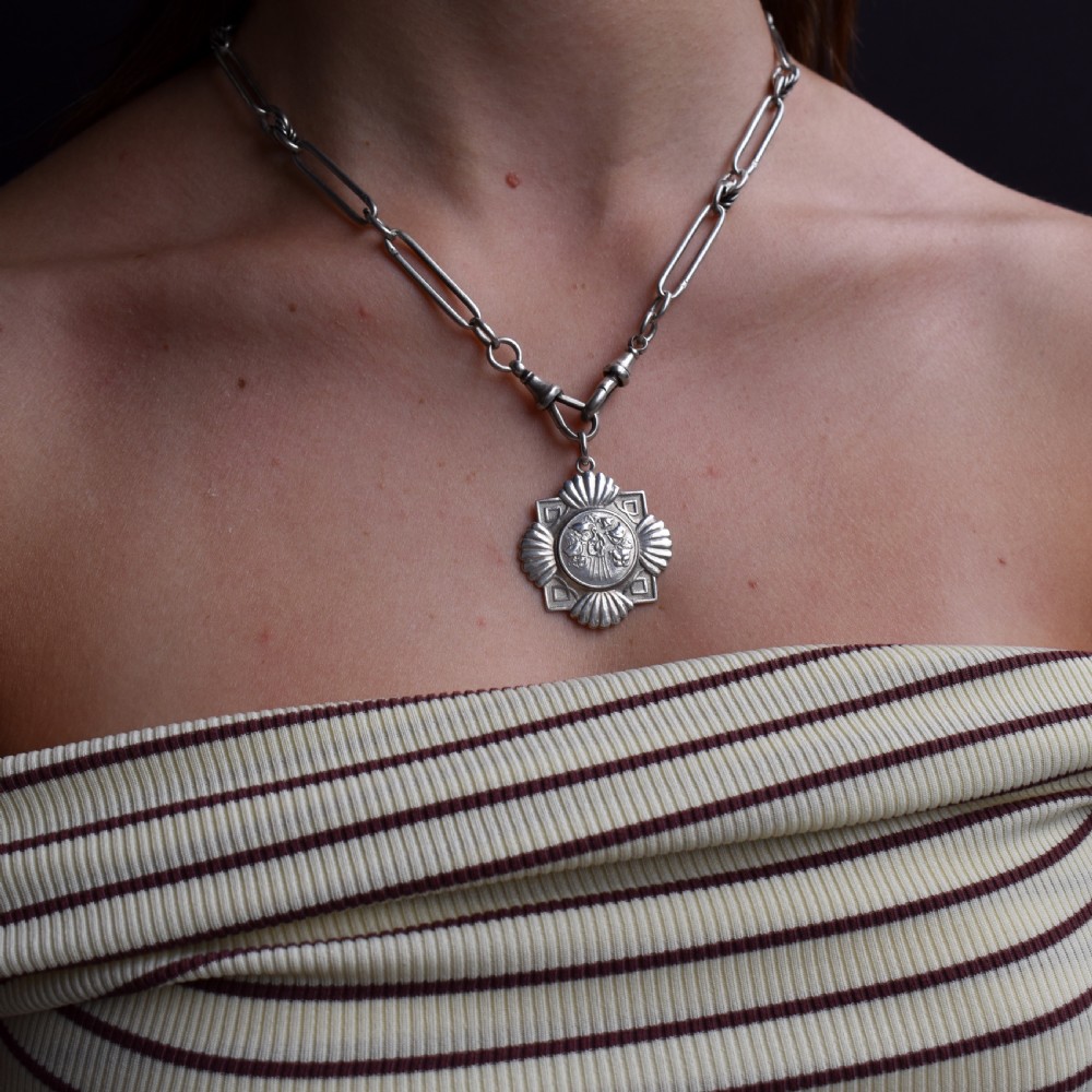 An Art Deco silver medallion featuring a central circular floral relief surrounded by a geometric square and fan-shaped fluted edges with a top suspension loop. View VIII