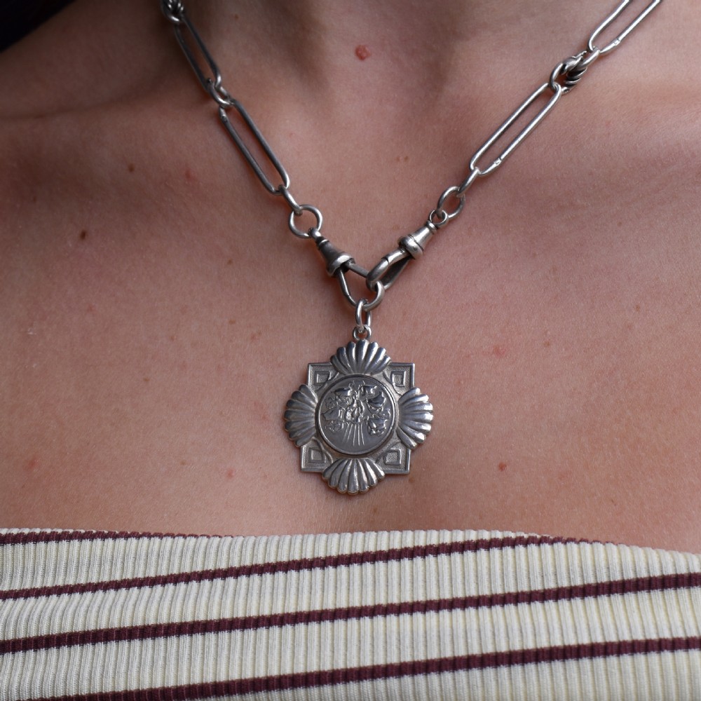An Art Deco silver medallion featuring a central circular floral relief surrounded by a geometric square and fan-shaped fluted edges with a top suspension loop. View IX