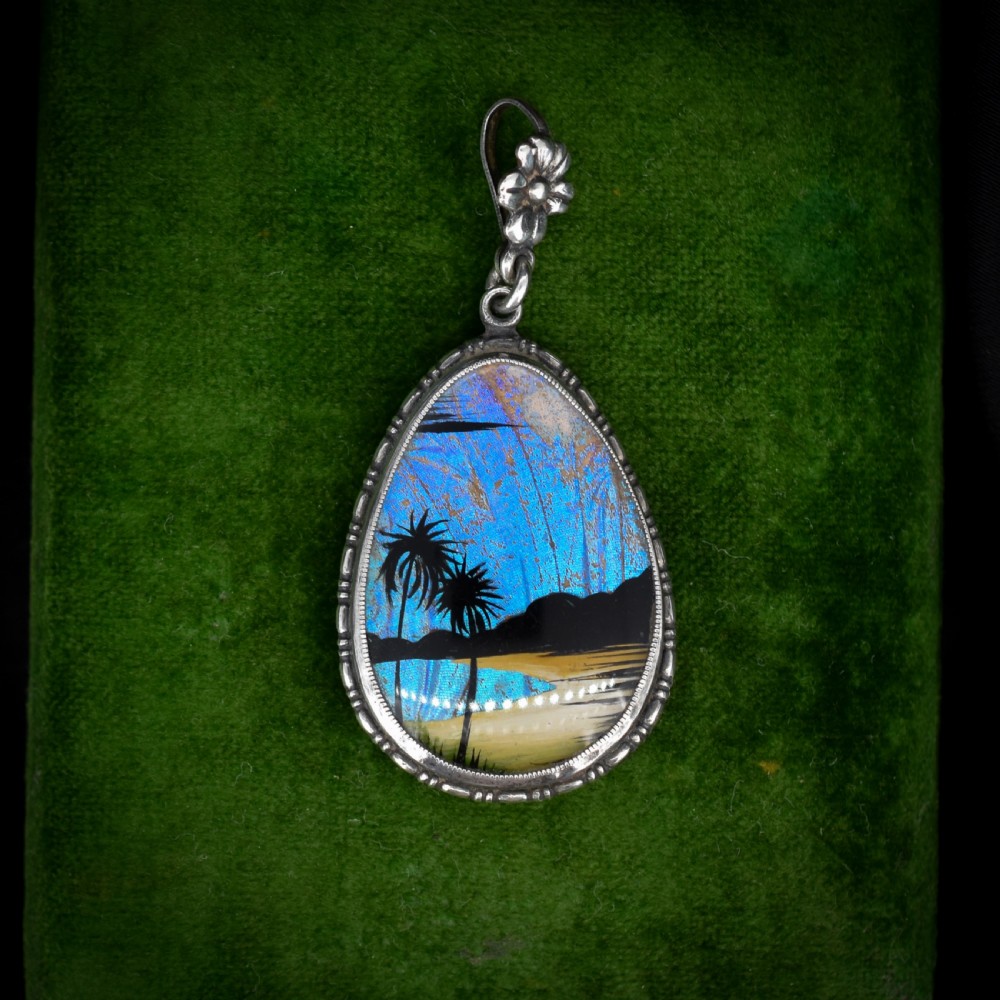 An Art Deco teardrop-shaped sterling silver pendant featuring a reverse-painted tropical palm scene over an iridescent blue butterfly wing.
