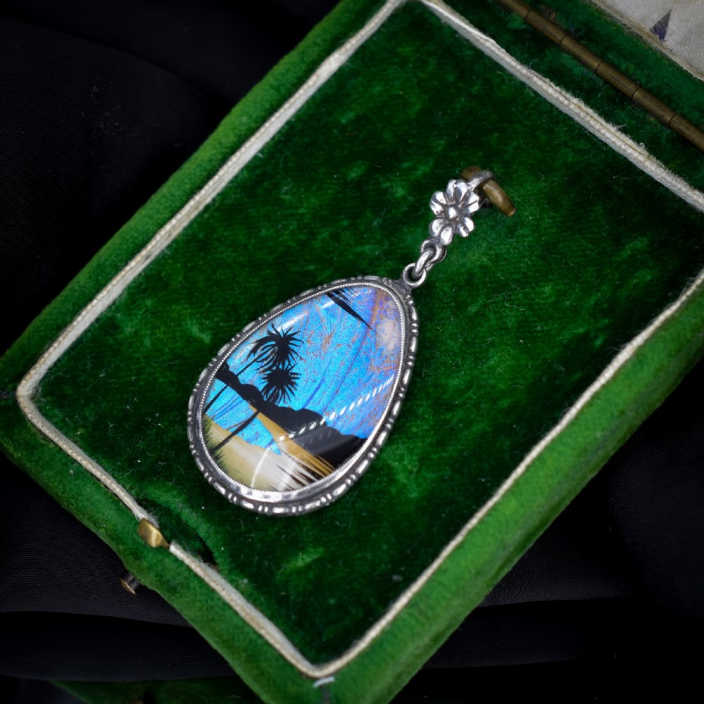An Art Deco teardrop-shaped sterling silver pendant featuring a reverse-painted tropical palm scene over an iridescent blue butterfly wing. View V