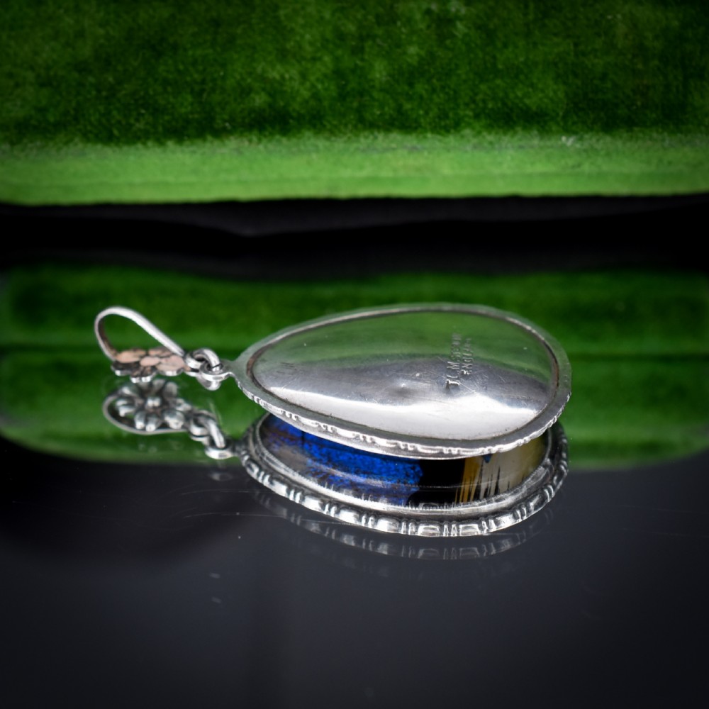 An Art Deco teardrop-shaped sterling silver pendant featuring a reverse-painted tropical palm scene over an iridescent blue butterfly wing. View X