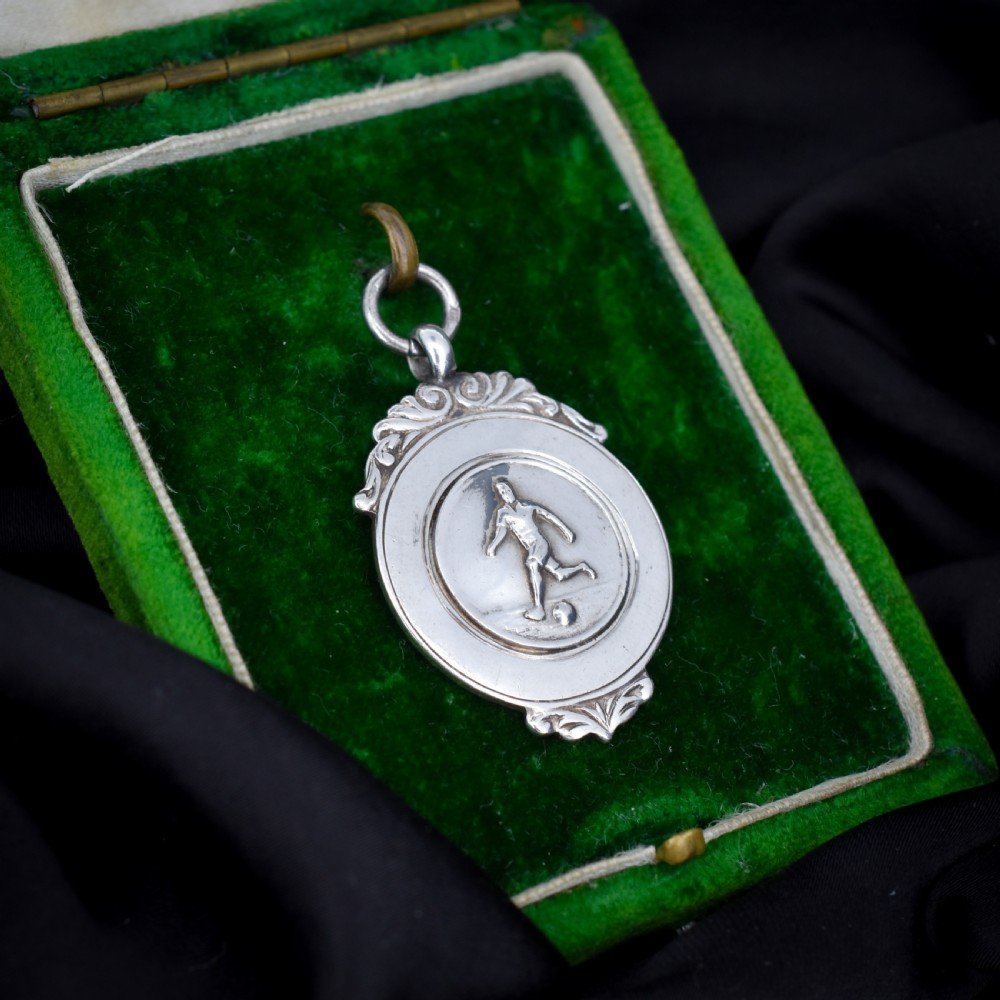 A circular silver Art Deco football medal pendant featuring an embossed figure of a footballer with a ball, framed by scrollwork edges and a top suspension ring. View II