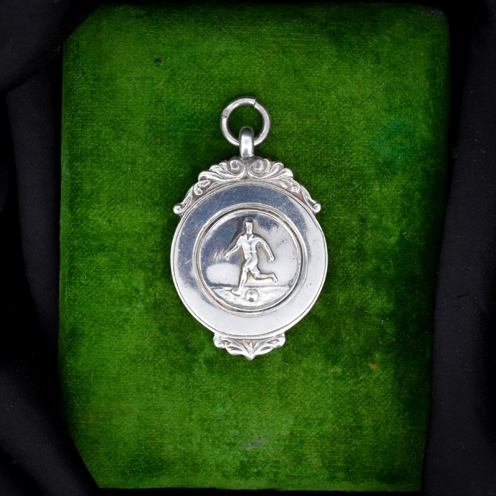 A circular silver Art Deco football medal pendant featuring an embossed figure of a footballer with a ball, framed by scrollwork edges and a top suspension ring. View III