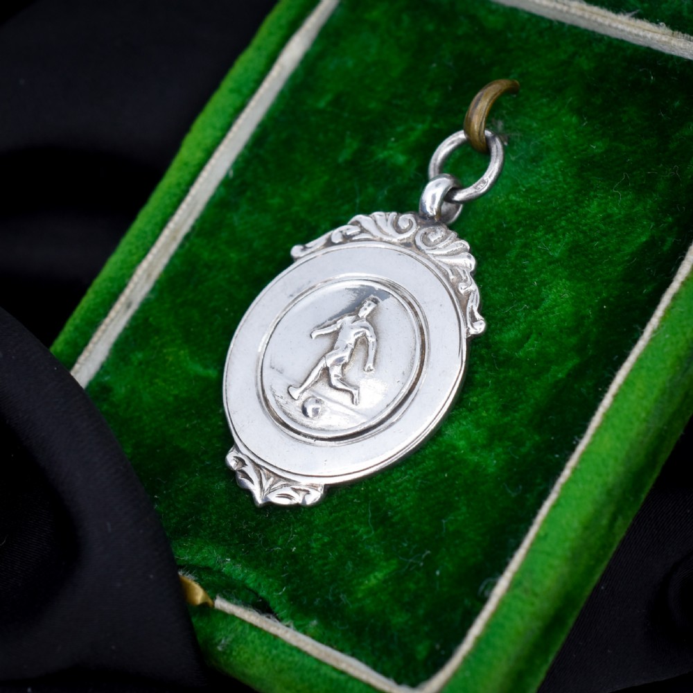A circular silver Art Deco football medal pendant featuring an embossed figure of a footballer with a ball, framed by scrollwork edges and a top suspension ring. View V