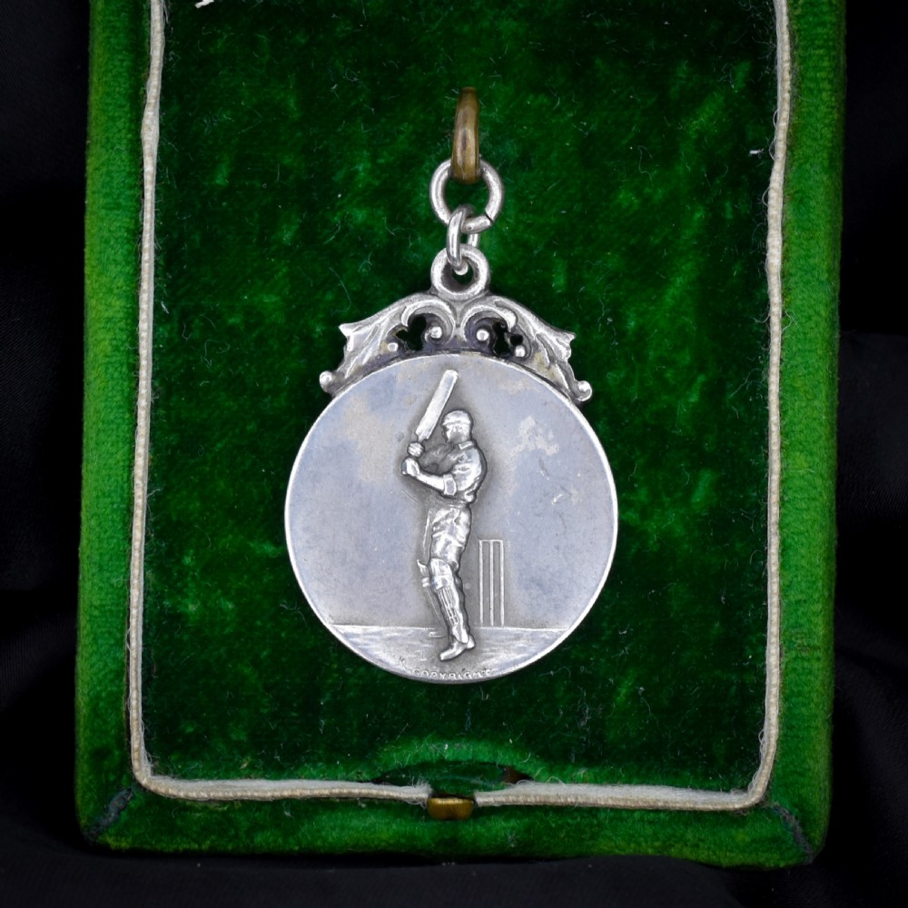 A circular silver Art Deco medallion depicting a raised relief figure of a cricketer with a bat beside wickets, finished with a scroll suspension loop. View II