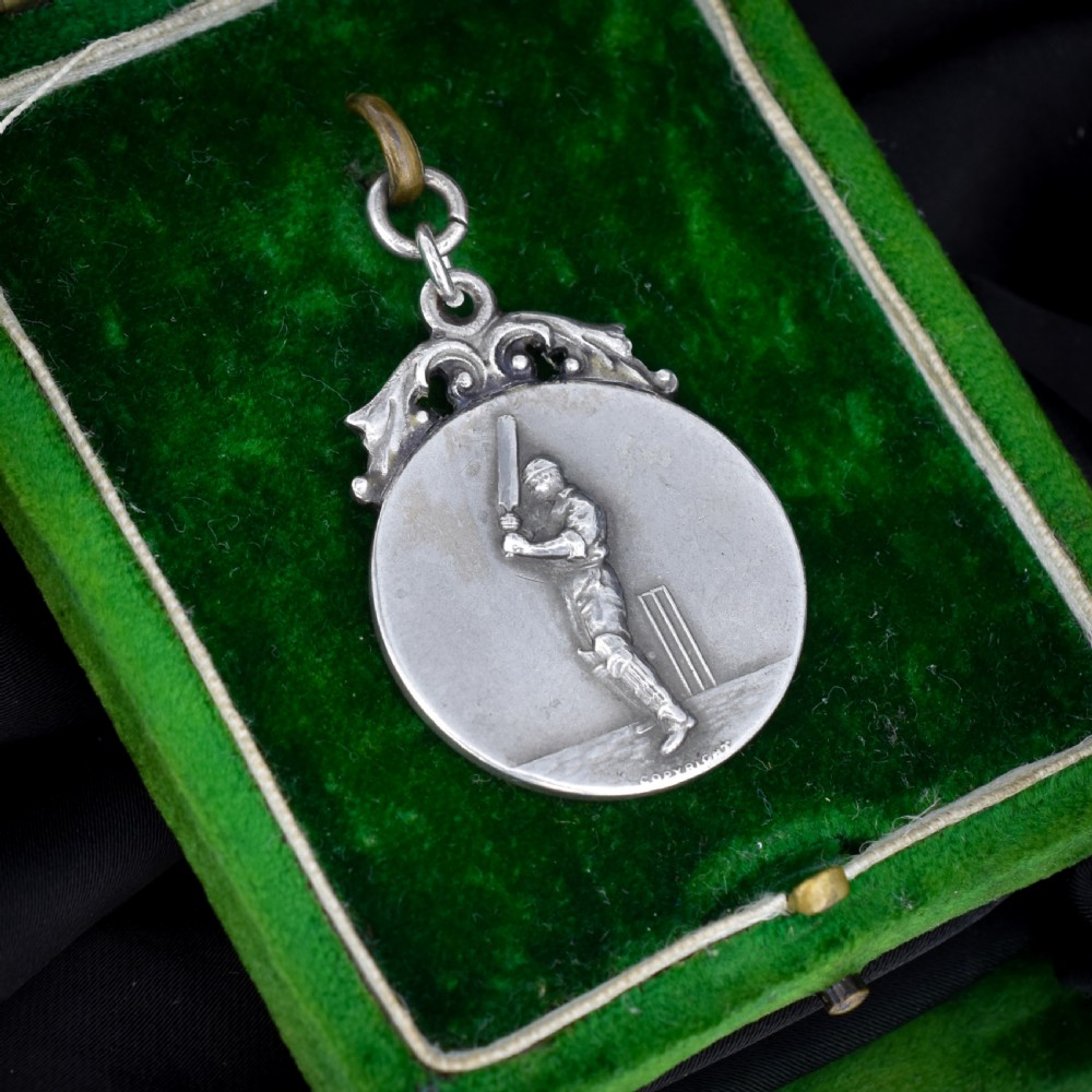 A circular silver Art Deco medallion depicting a raised relief figure of a cricketer with a bat beside wickets, finished with a scroll suspension loop. View III