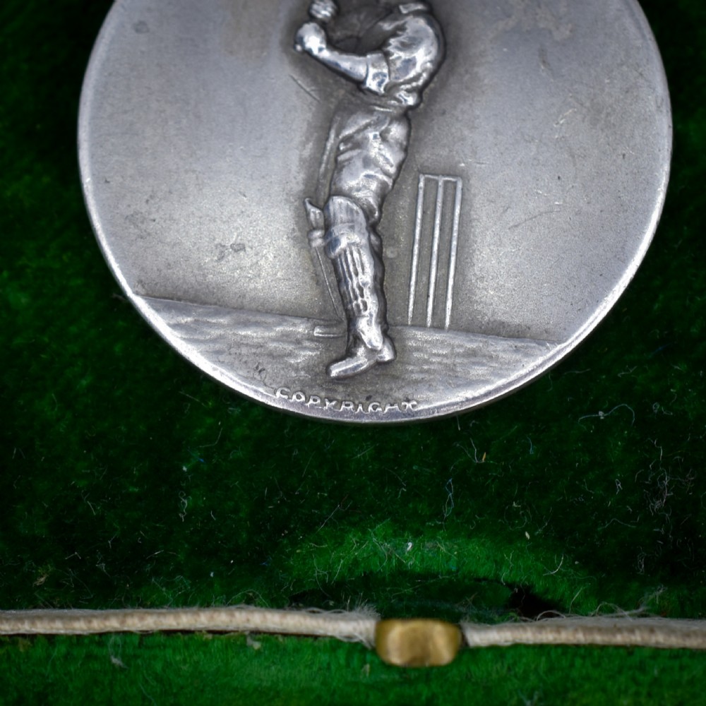 A circular silver Art Deco medallion depicting a raised relief figure of a cricketer with a bat beside wickets, finished with a scroll suspension loop. View IV