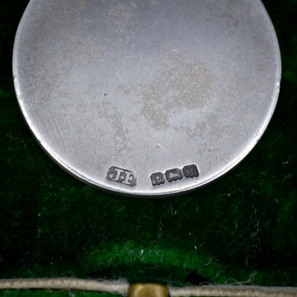 A circular silver Art Deco medallion depicting a raised relief figure of a cricketer with a bat beside wickets, finished with a scroll suspension loop. View V