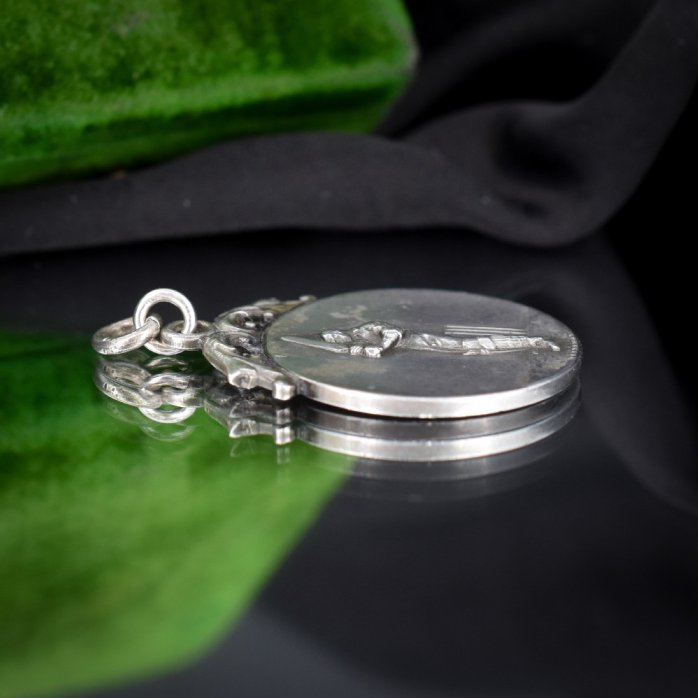 A circular silver Art Deco medallion depicting a raised relief figure of a cricketer with a bat beside wickets, finished with a scroll suspension loop. View VII