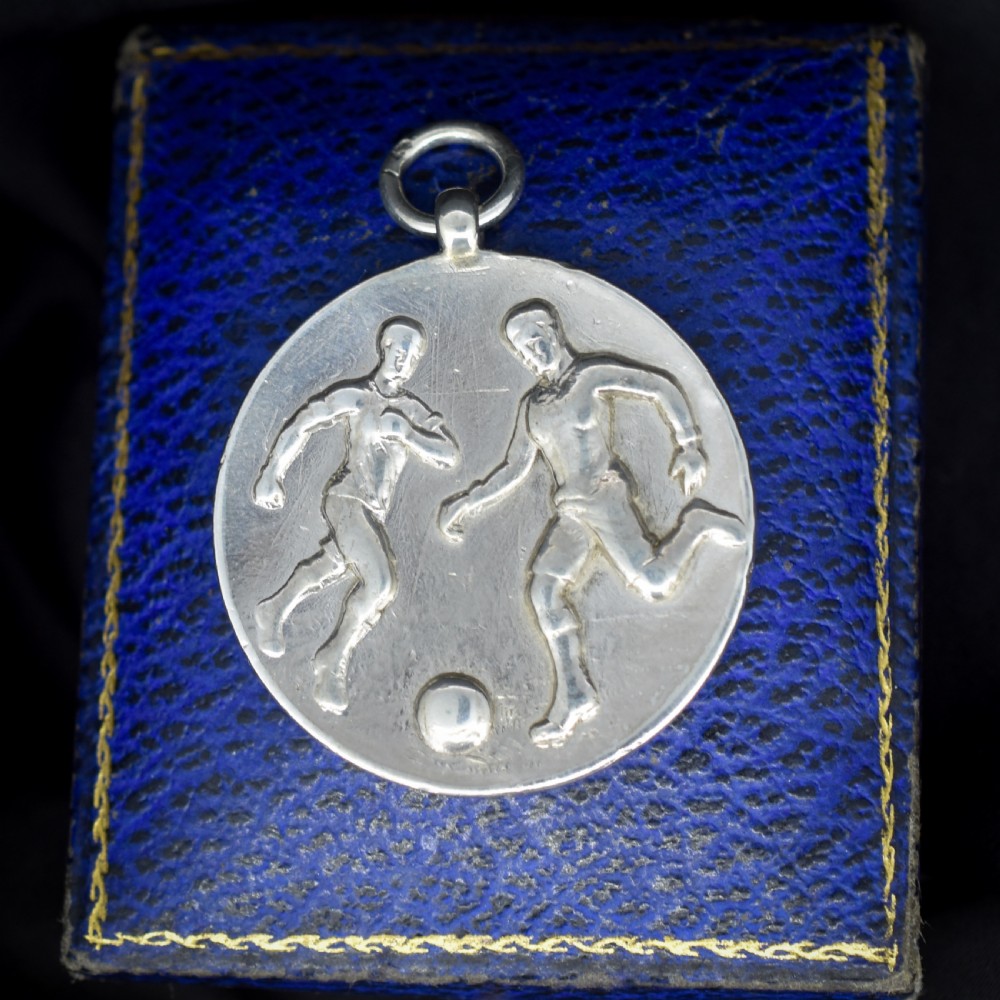 A circular silver Art Deco medallion depicting two football players in relief with a football, suspended from a top loop.