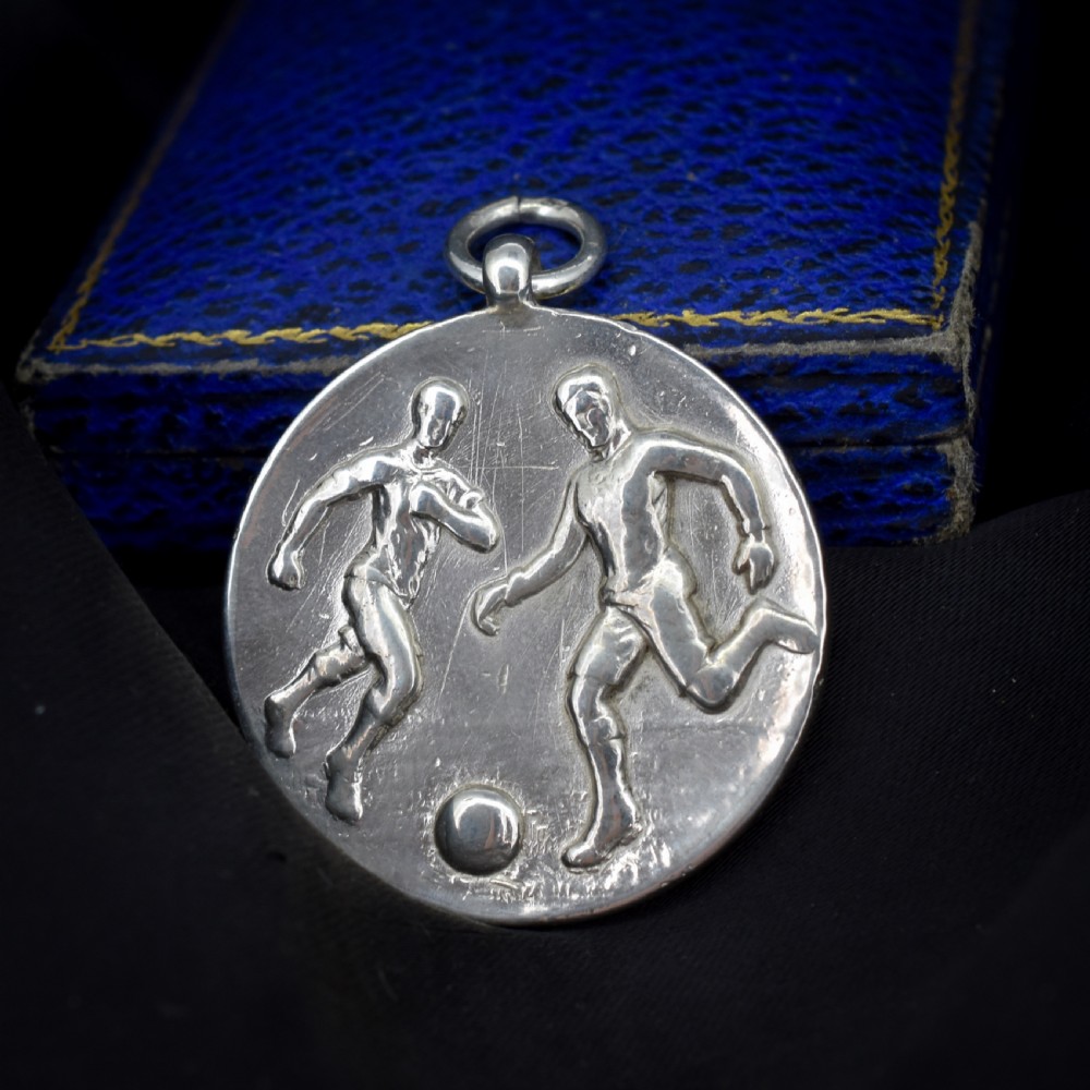 A circular silver Art Deco medallion depicting two football players in relief with a football, suspended from a top loop. View II