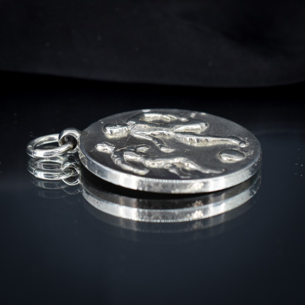 A circular silver Art Deco medallion depicting two football players in relief with a football, suspended from a top loop. View III