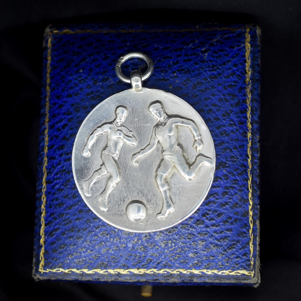 A circular silver Art Deco medallion depicting two football players in relief with a football, suspended from a top loop. View V