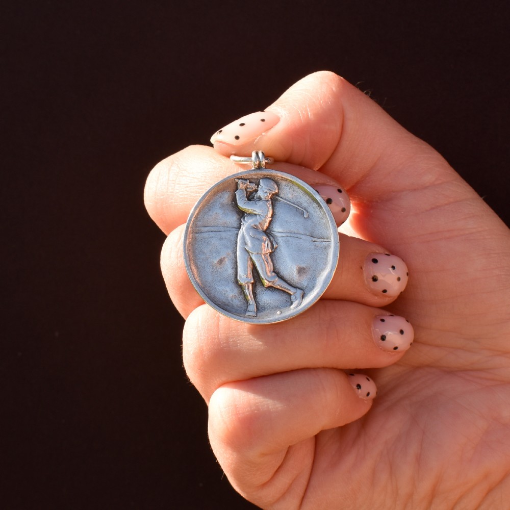 A circular silver medallion featuring a golfer rendered in relief mid-swing with a suspension ring attached to the top. View IX