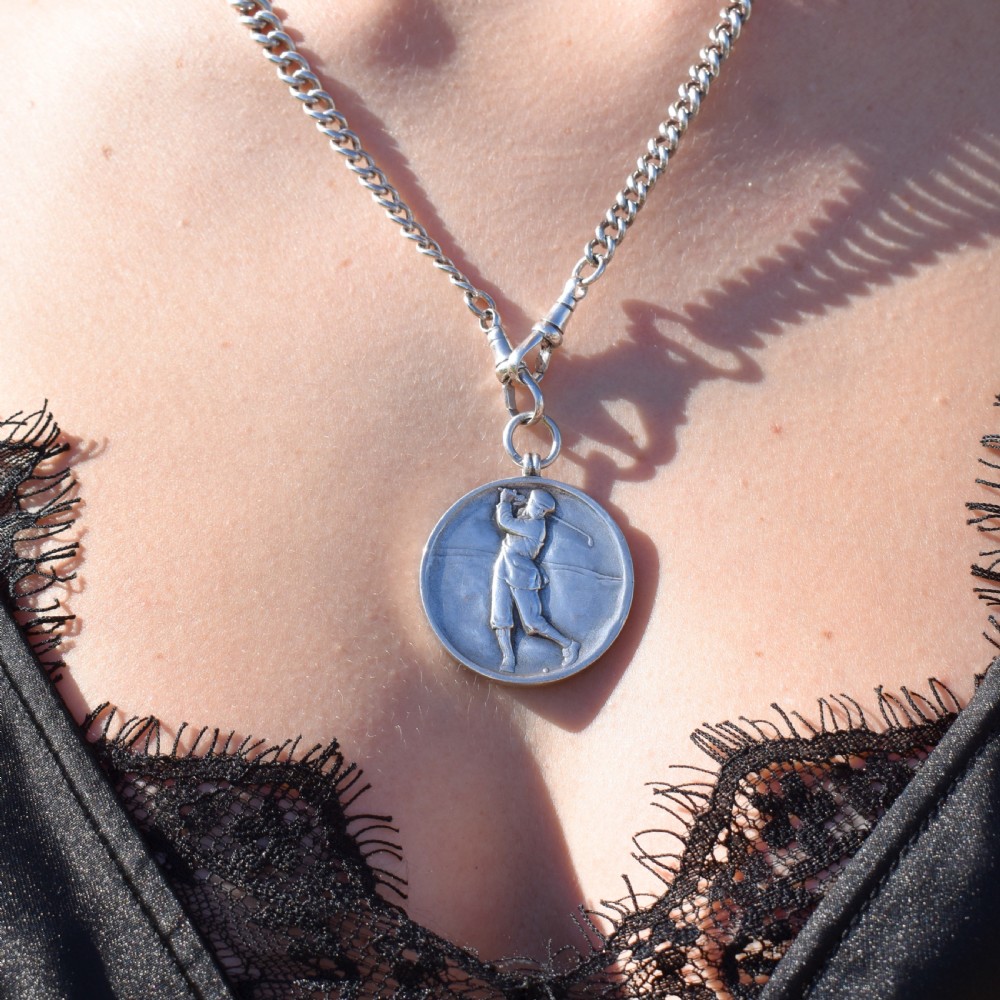 A circular silver medallion featuring a golfer rendered in relief mid-swing with a suspension ring attached to the top. View X