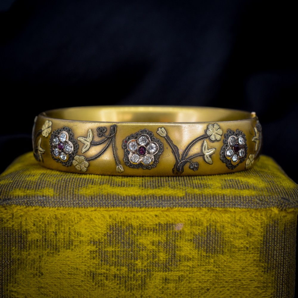 An antique gold-filled hinged bangle decorated with raised floral motifs with red and clear paste stone centres.