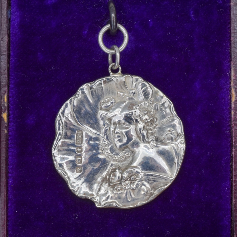 A circular Art Nouveau sterling silver pendant featuring a relief portrait of a woman surrounded by floral motifs and hallmarked on the front surface.
