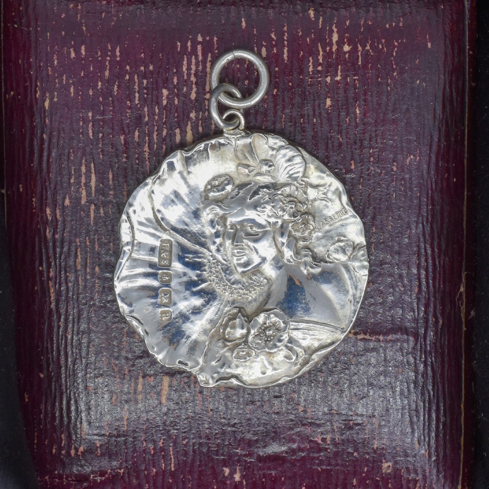 A circular Art Nouveau sterling silver pendant featuring a relief portrait of a woman surrounded by floral motifs and hallmarked on the front surface. View II