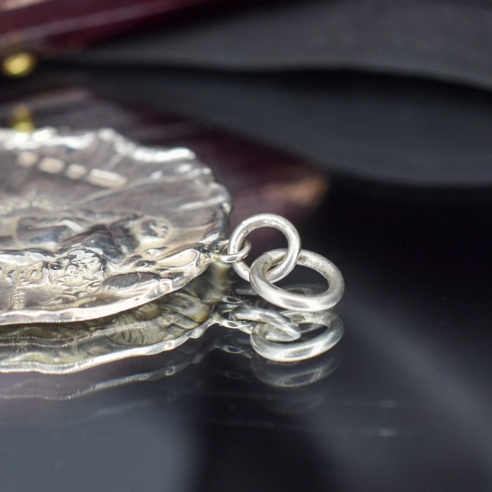 A circular Art Nouveau sterling silver pendant featuring a relief portrait of a woman surrounded by floral motifs and hallmarked on the front surface. View IX