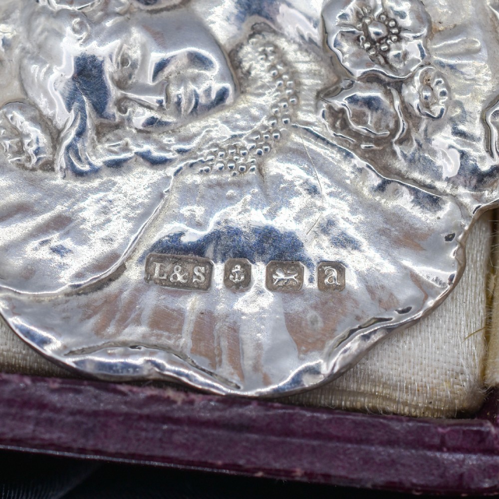 A circular Art Nouveau sterling silver pendant featuring a relief portrait of a woman surrounded by floral motifs and hallmarked on the front surface. View X