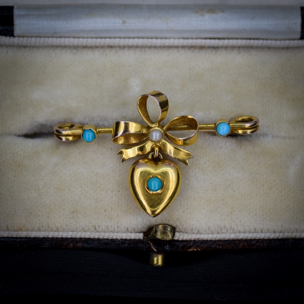 An antique yellow gold bar brooch featuring a central bow motif with a seed pearl, a dangling gold heart set with a turquoise cabochon, and two flanking turquoise stones.