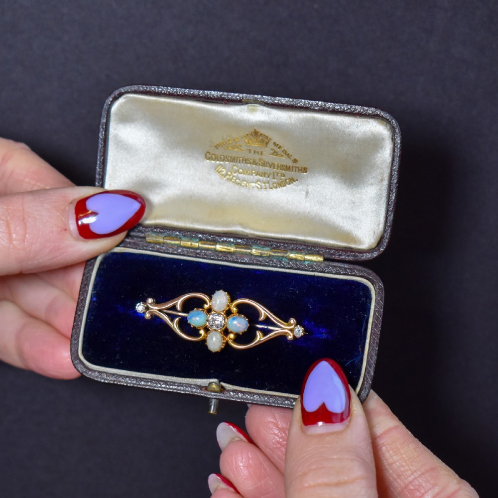 An antique 15ct gold brooch featuring four cabochon opals and central diamond accents set in an openwork scroll framing, contained inside a navy velvet presentation box.