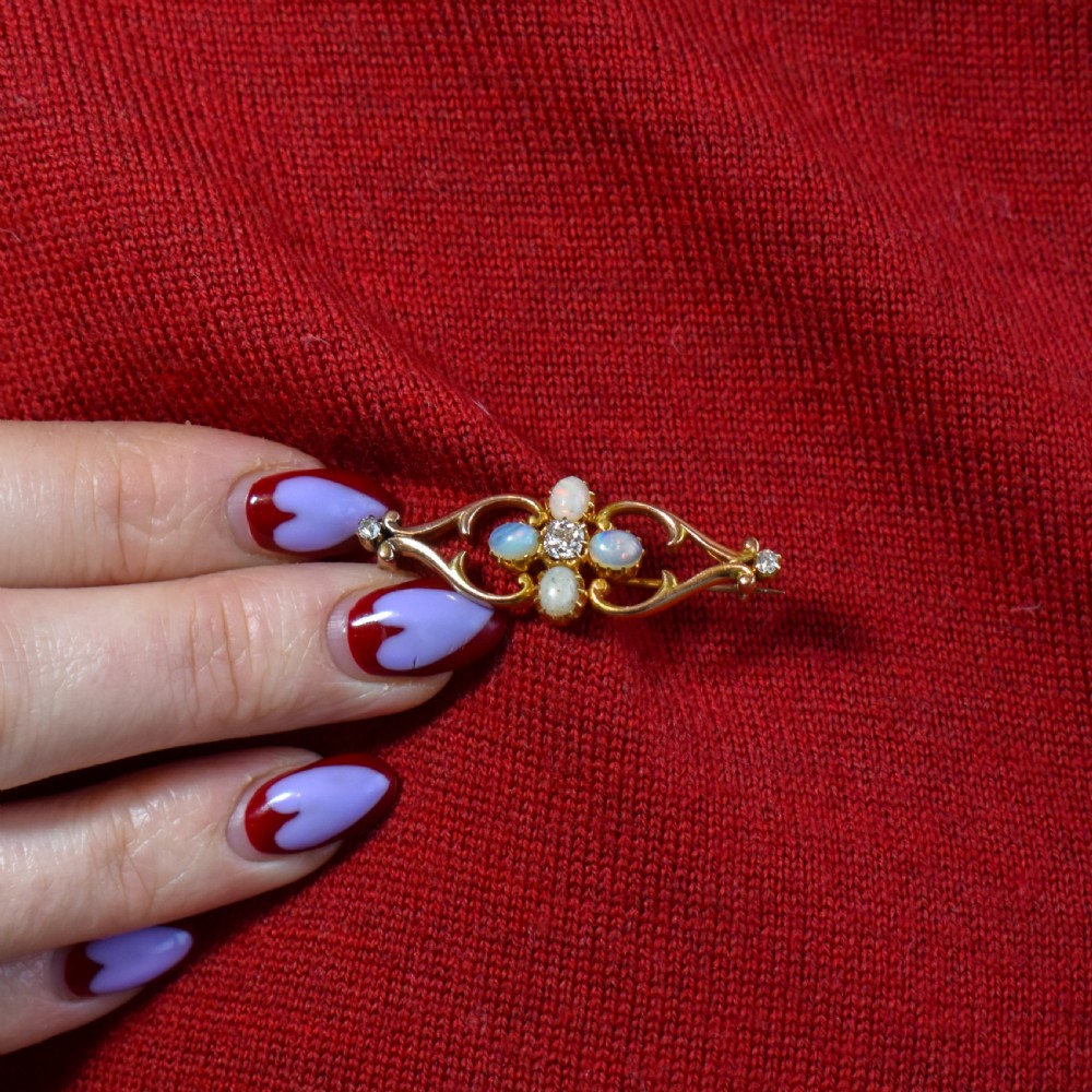 An antique 15ct gold brooch featuring four cabochon opals and central diamond accents set in an openwork scroll framing, contained inside a navy velvet presentation box. View V