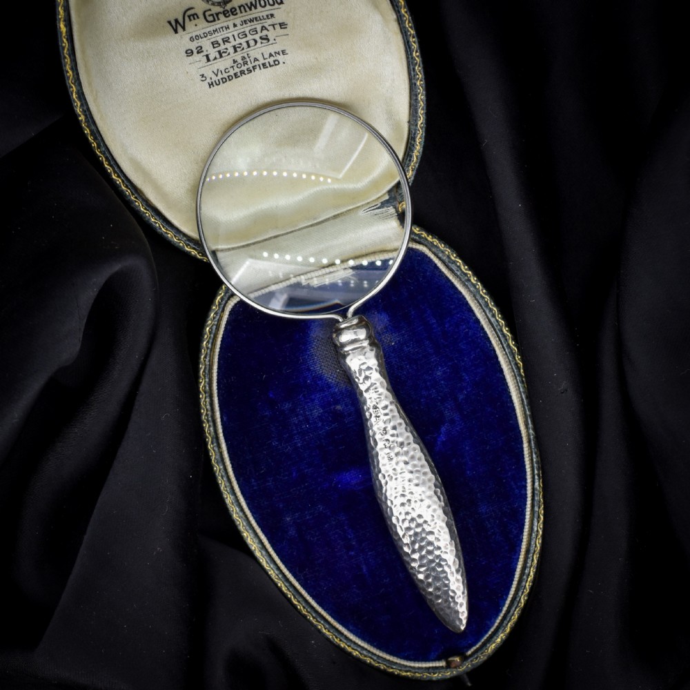 An Edwardian sterling silver magnifying glass featuring a hammered planished handle and a circular glass lens, presented inside an open velvet-lined case. View V