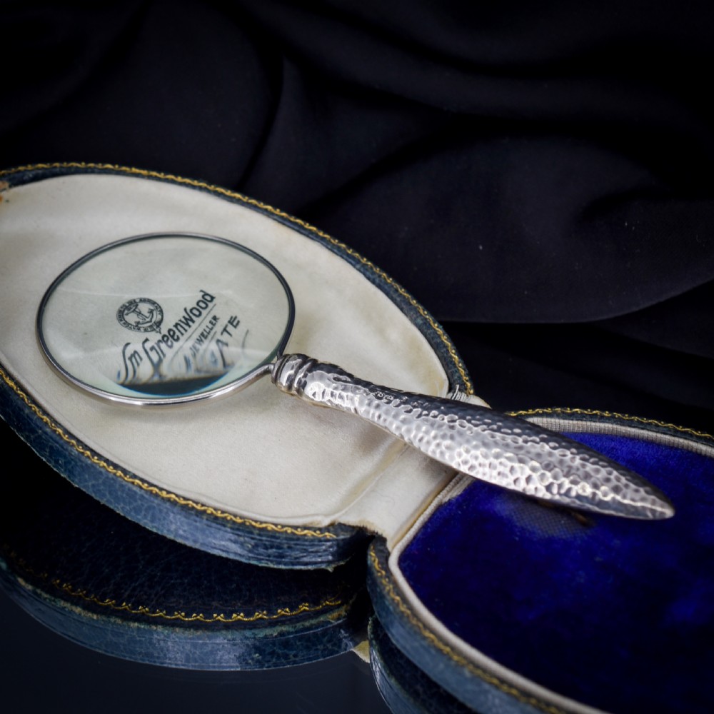 An Edwardian sterling silver magnifying glass featuring a hammered planished handle and a circular glass lens, presented inside an open velvet-lined case. View VI