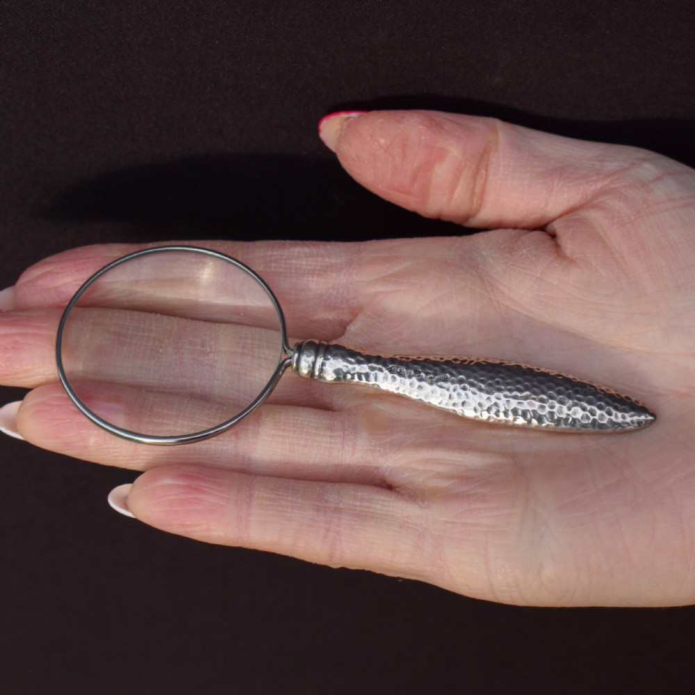 An Edwardian sterling silver magnifying glass featuring a hammered planished handle and a circular glass lens, presented inside an open velvet-lined case. View VII