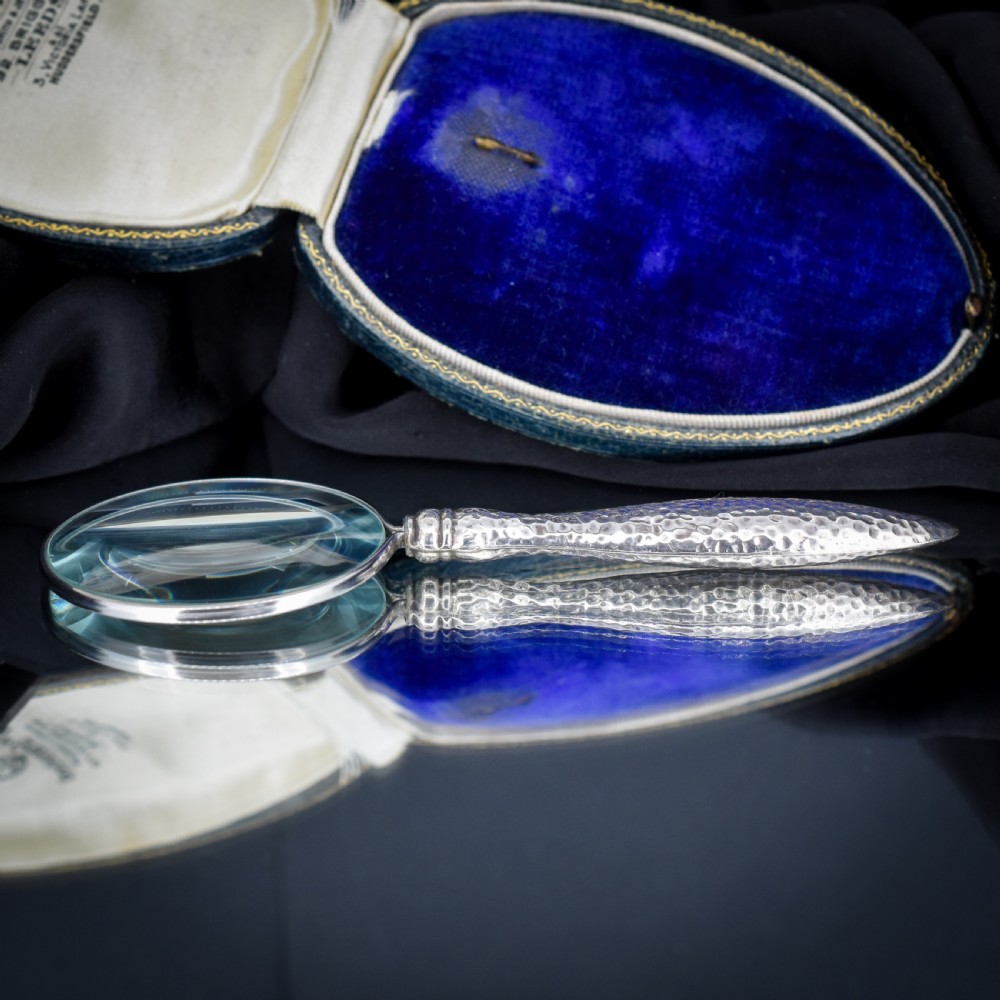 An Edwardian sterling silver magnifying glass featuring a hammered planished handle and a circular glass lens, presented inside an open velvet-lined case. View X