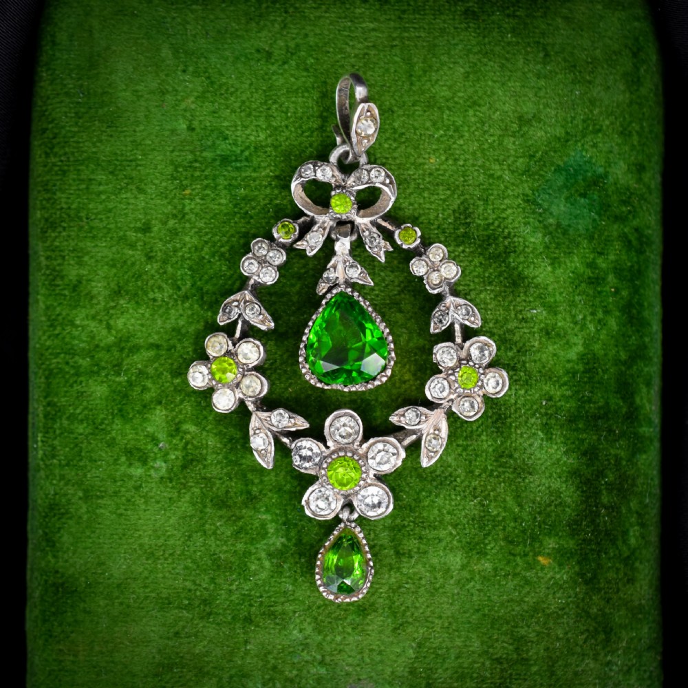 An Edwardian silver openwork floral lavalier pendant set with clear and green paste glass stones featuring a bow motif and suspended green teardrop stone.
