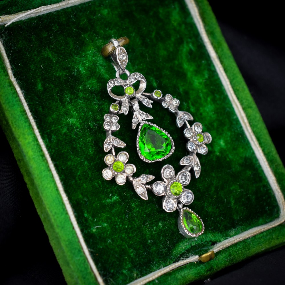 An Edwardian silver openwork floral lavalier pendant set with clear and green paste glass stones featuring a bow motif and suspended green teardrop stone. View V