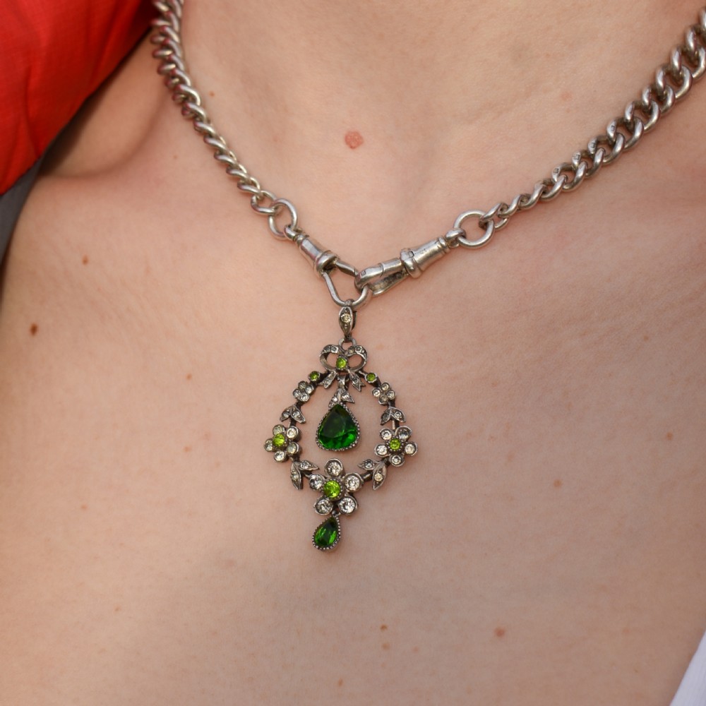 An Edwardian silver openwork floral lavalier pendant set with clear and green paste glass stones featuring a bow motif and suspended green teardrop stone. View VI