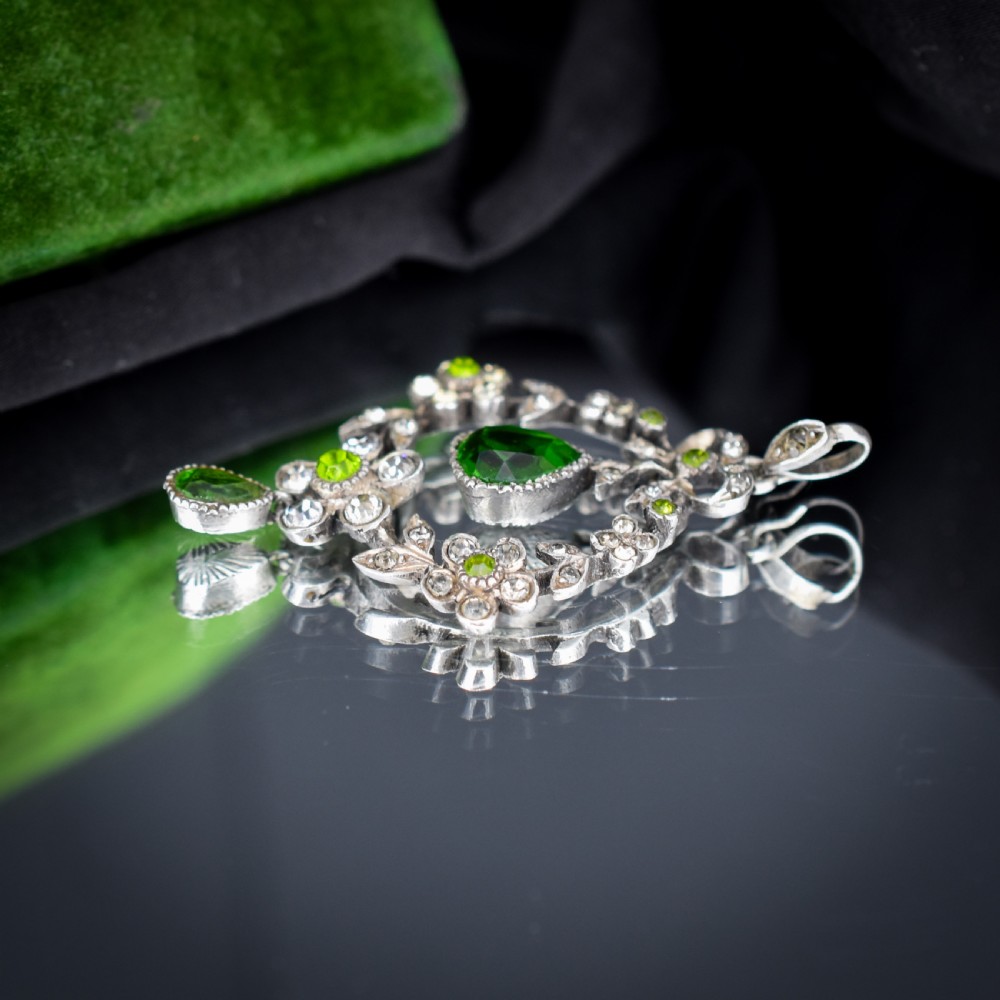 An Edwardian silver openwork floral lavalier pendant set with clear and green paste glass stones featuring a bow motif and suspended green teardrop stone. View VII