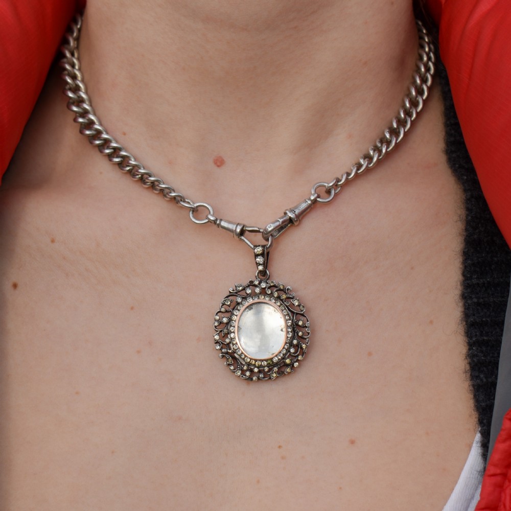 An oval antique locket pendant crafted in silver and gold, featuring an openwork scrollwork border set with old-cut clear paste stones surrounding a central glass compartment. View II