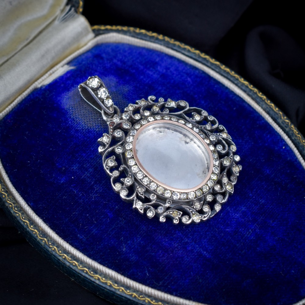 An oval antique locket pendant crafted in silver and gold, featuring an openwork scrollwork border set with old-cut clear paste stones surrounding a central glass compartment. View III